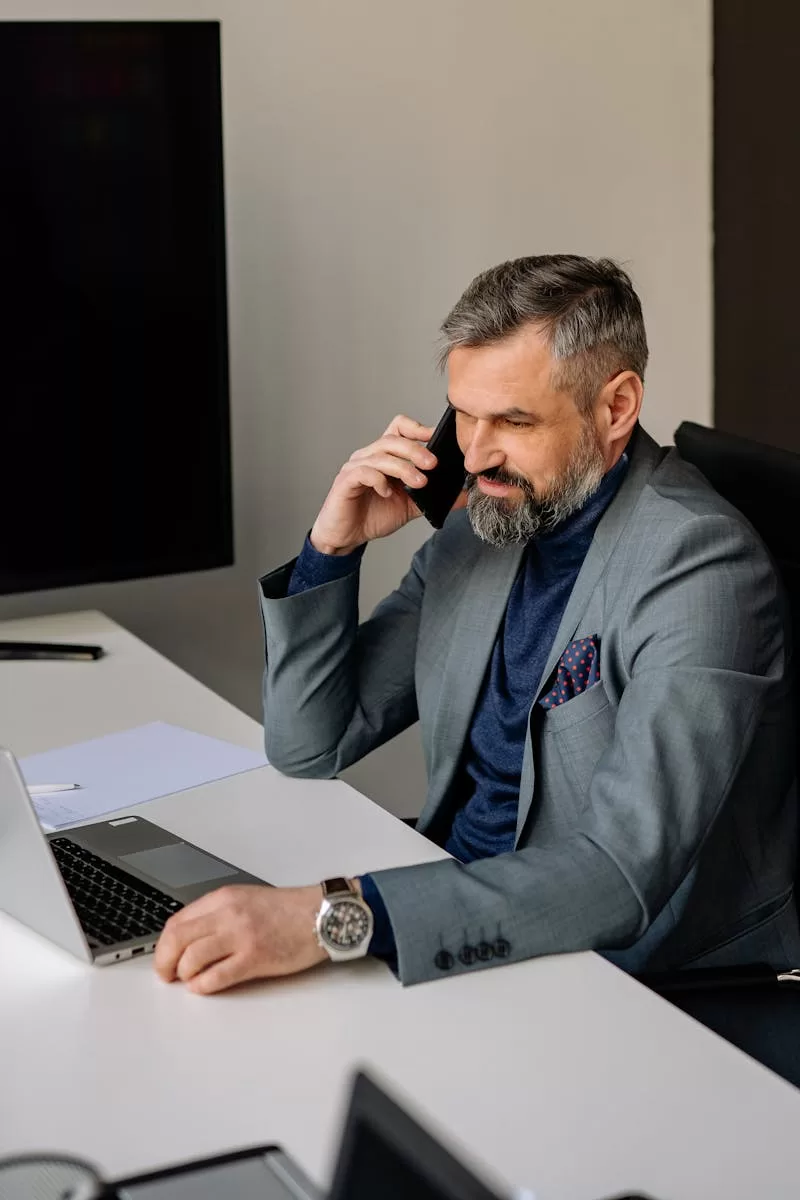 Bearded businessman in office using laptop and phone, dressed in formal attire.